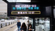 Commuters stand at a bus stop with an electronic display that reads 