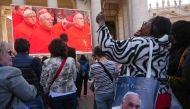 An attendee wearing a bag depicting the late Pope Francis as she watches a giant screen displaying images of the holy mass of cardinals to the Sistine chapel, at St Peter's Square on the first day of the conclave to elect the next pope, at the Vatican, on May 7, 2025. (Photo by Dimitar DILKOFF / AFP)