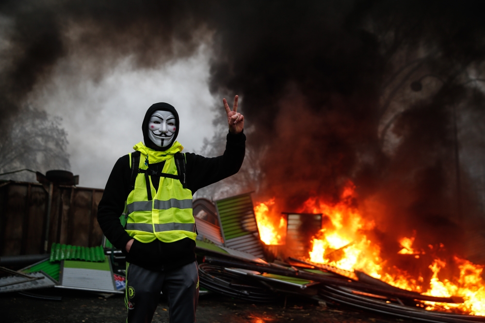A protester wearing a Guy Fawkes mask makes the victory sign near a burning barricade during a protest of Yellow vests (Gilets jaunes) against rising oil prices and living costs, on December 1, 2018 in Paris. / AFP / Abdulmonam EASSA