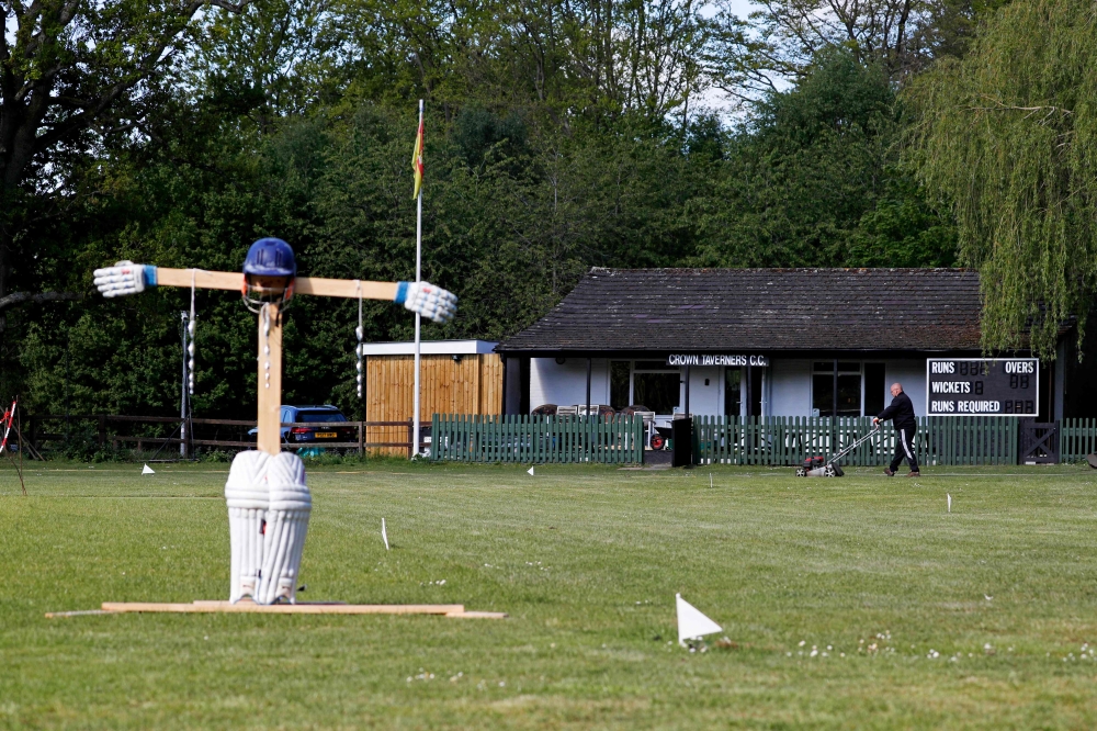 Gary O'Hara, volunteer groundsman and semi-retired player for Crown Taverners in the Hampshire cricket league, mows the grass on the ground in front of the club house with a cricket-themed scarecrow standing sentinal at Crown Taverners Cricket Club in Cam