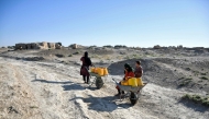 An Afghan woman with children takes a break as they haul water cans on wheelbarrows along a deserted street in the drought-ridden village of Bolak at Chahar Bolak district, Balkh province on July 10, 2025.  (Photo by Atif Aryan / AFP)
