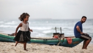 A displaced Palestinian girl runs along the beach as a man sits on a fishing boat, in Deir el-Balah in the central Gaza Strip, on October 5, 2025. (Photo by Bashar TALEB / AFP)
