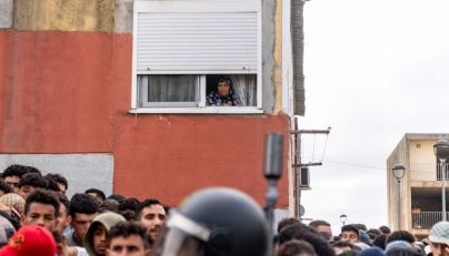 A resident watches from her window migrants waiting on the sidewalk before being registered in Loma Colmenar district of Ceuta on August 31, 2026. Photo by Antonio Sempere / AFP