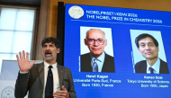 The chair of the Nobel Committee for Chemistry Heiner Linke (L) explains the field of research of Nobel Prize winners France's Henri Kagan and Japan's Kenso Soai (displayed on the screen in background) during a press conference in Stockholm, Sweden, on October 7, 2026. (Photo by Jonathan Nackstrand / AFP)