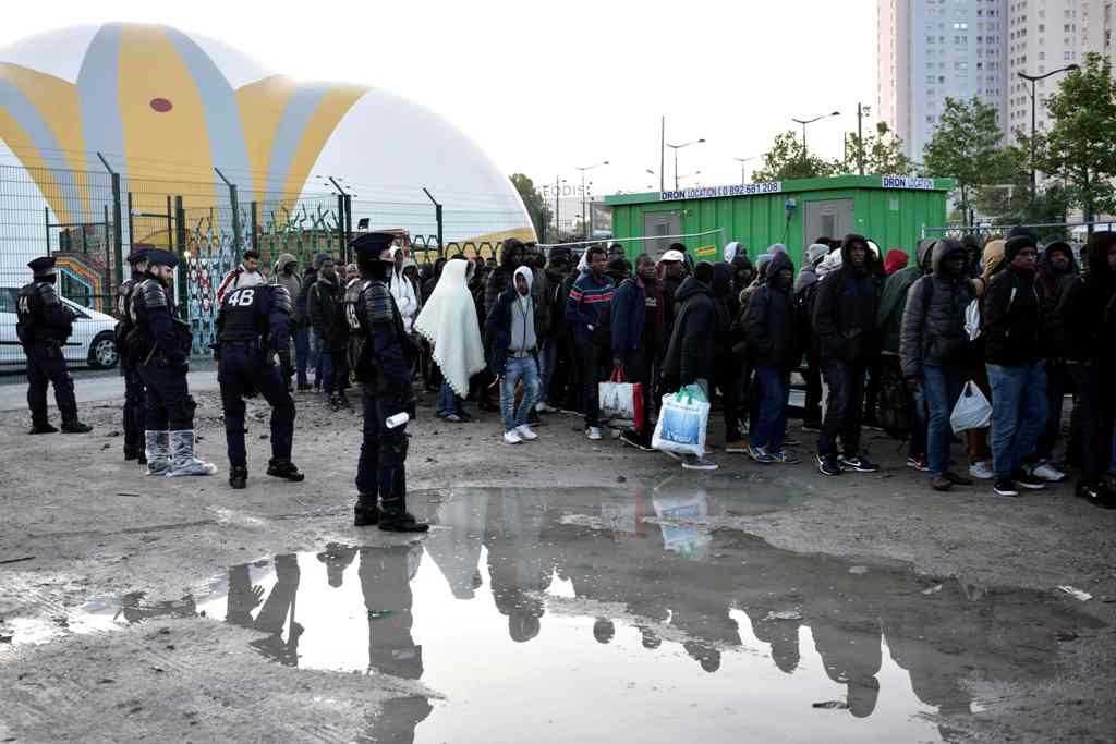 French anti-riot police CRS officers stand guard by migrants and refugees queueing by the migrant camp at Porte de la Chapelle during the evacuation of a makeshift camp near La Chapelle, in Paris, on May 9, 2017, one of several camps sprouting up around t