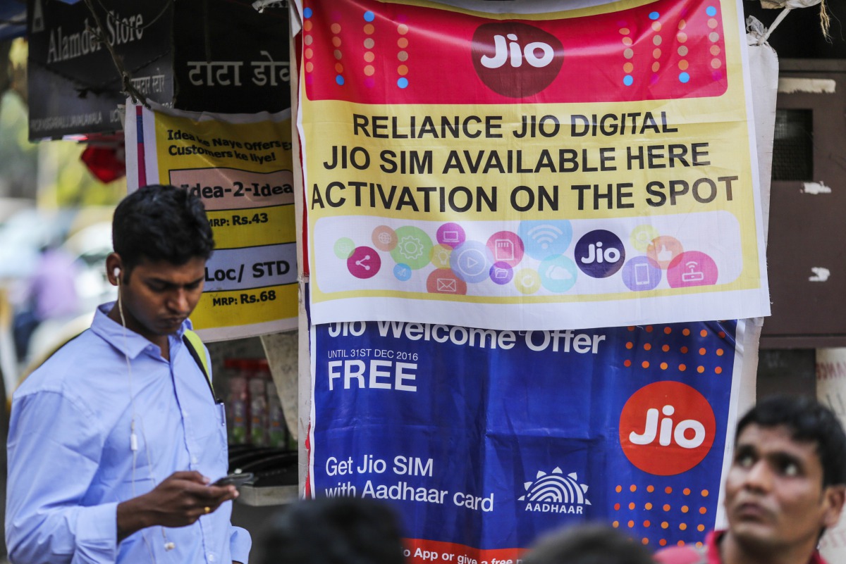 A pedestrian using a mobile phone walks past banners for Reliance Jio, the mobile network of Reliance Industries Ltd., and Bharti Airtel Ltd. outside a mobile phone store in Mumbai, India, on Oct. 24, 2016.(Bloomberg photo by Dhiraj Singh)