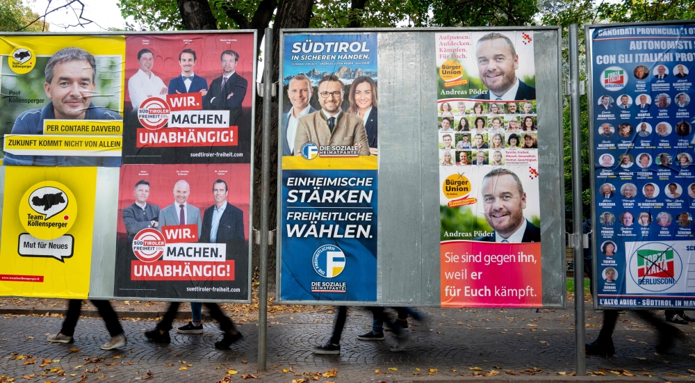 People pass behind election posters in Bolzano, Italy on October 15, 2018. AFP / Joe Klamar 
