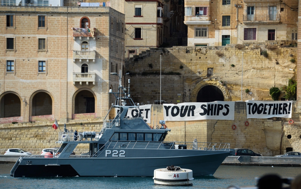 A Malta Coast Guards ship patrols as protesters display a banner reading 