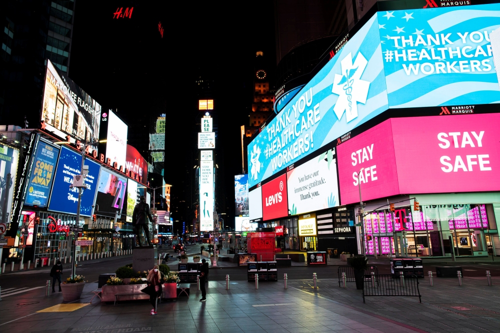 People walk in Times Square, Manhattan while some screens are seen illuminated in blue as part of the 