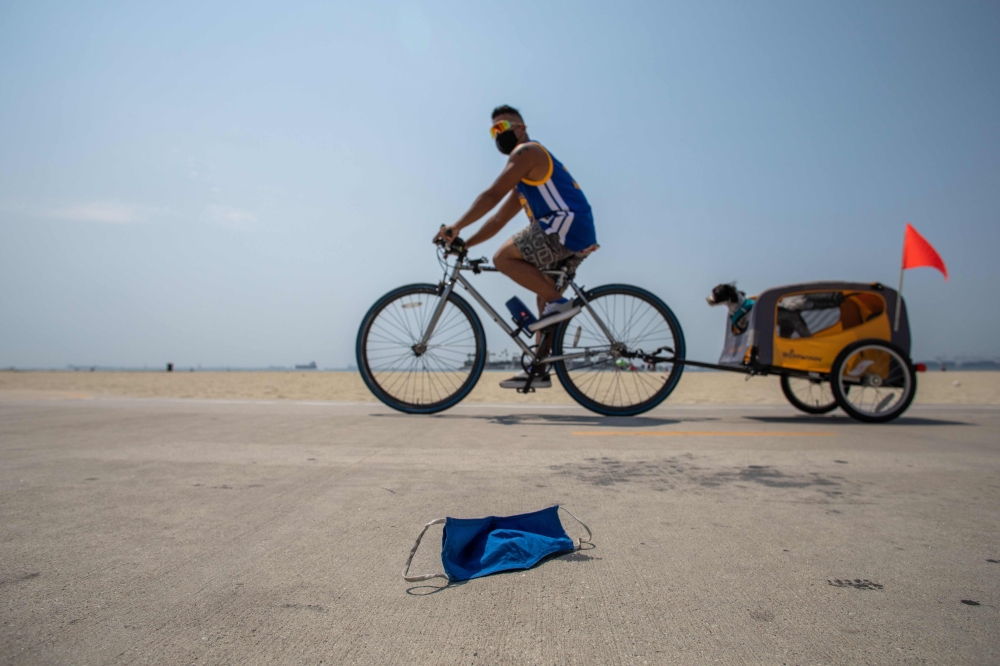A man wearing face mask rides his bike towing a dog next to a discarded face mask in the bike path in Long Beach, California on August 22, 2020.  AFP / Apu GOMES
