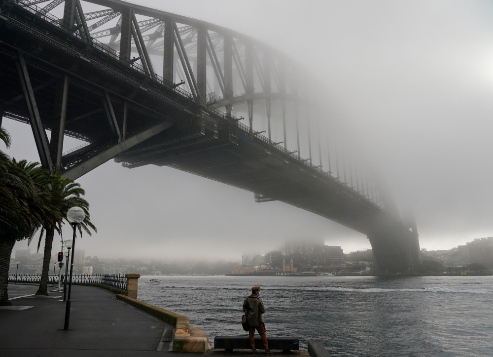 A man wearing a protective face mask takes in the waterfront view underneath the Sydney Harbour Bridge, seen shrouded in fog, during a lockdown to curb the spread of a coronavirus disease (COVID-19) outbreak in Sydney, Australia, June 30, 2021. REUTERS/Lo