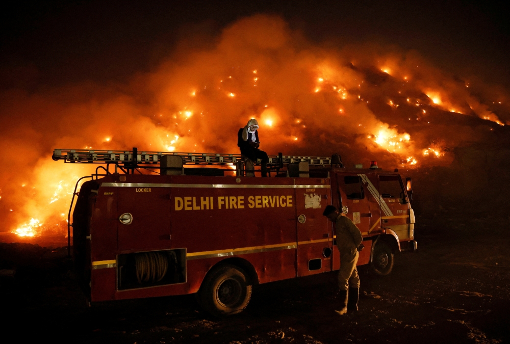 A firefighter uses his mobile phone as he sits on top of a fire truck as smoke billows from burning garbage at the Bhalswa landfill site in New Delhi, India, April 27, 2022. REUTERS/Adnan Abidi
