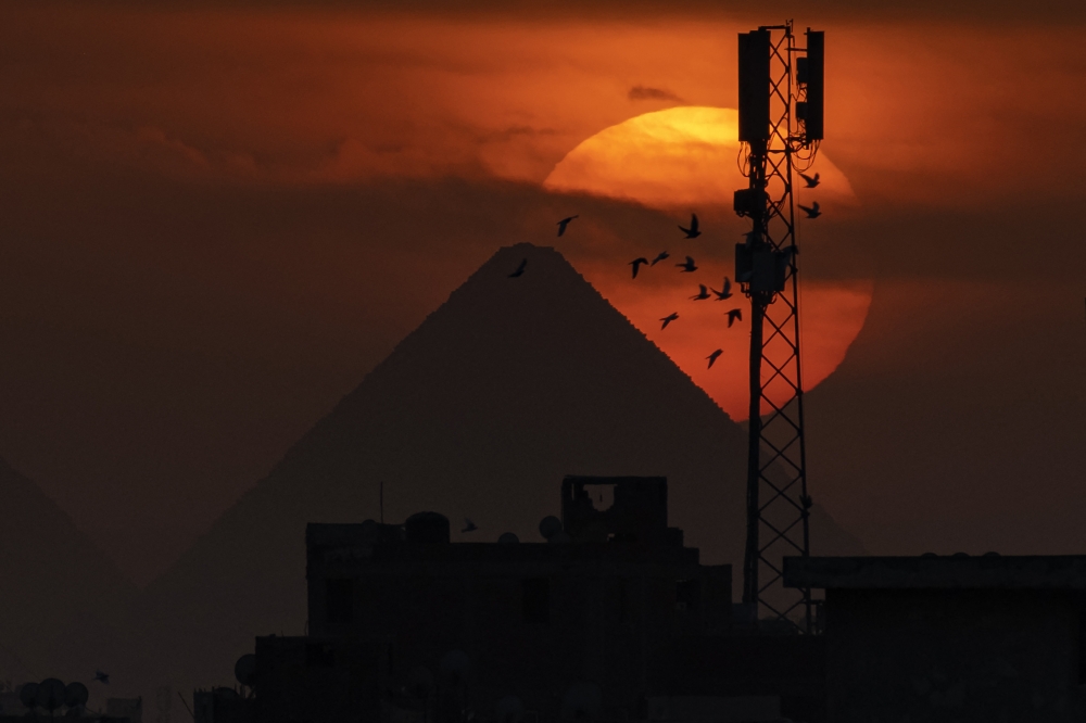 In this file photo taken on November 20, 2022, a flock of pigeons flies as the sun sets behind the Great Pyramid of Khufu and a cellular broadcast antenna tower in Giza, the twin city of Egypt's capital Cairo, on November 20, 2022. (Photo by Amir MAKAR / AFP)