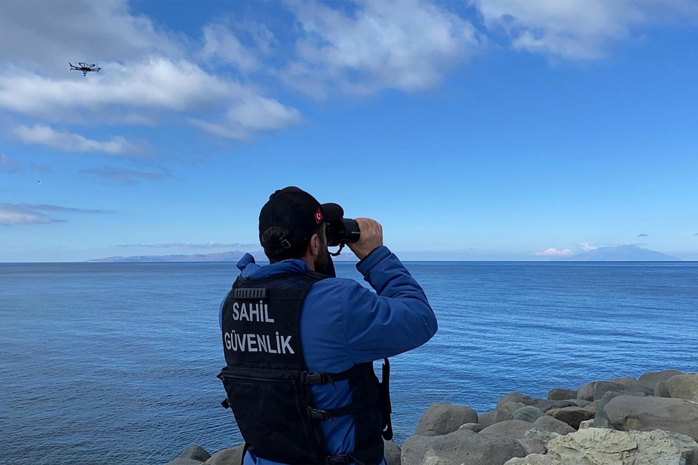 :This handout photograph taken and released on March 15, 2024 in Canakkale by Turkish news agency DHA (Demiroren News Agency) shows a coast guard officer looking through binoculars as a drone seaches for victims, after at least eight migrants drowned after a boat carrying them capsized off the Turkish coast in the Aegean Sea. (Photo by Demiroren News AAgency / DHA (Demiroren News Agency) / AFP)